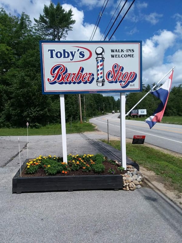 Toby's Barber Shop roadside sign on Route 25 in Meredith, New Hampshire, painted red white and blue with a barber pole and the words Walk-Ins Welcome